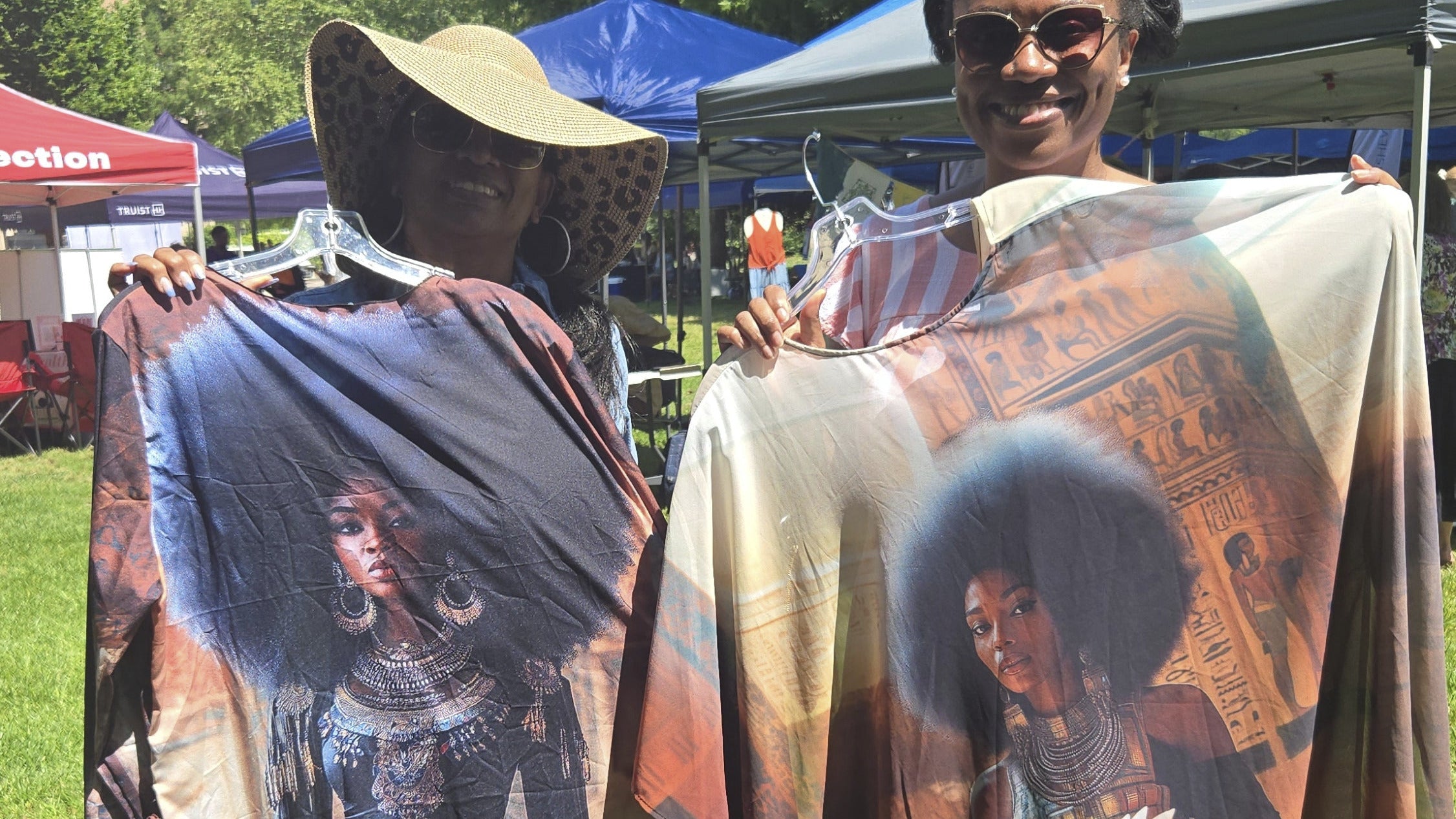 Mother and daughter holding up long robes just purchased AMNIBLACK depictions of women, one with an afro, at an outdoor event.